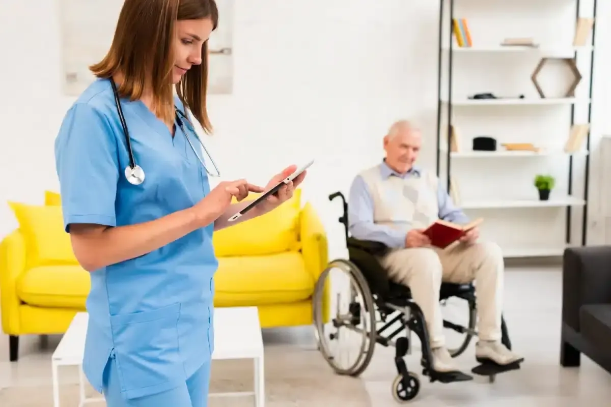 Enfermera de uniforme azul que usa una tableta, con un hombre mayor en silla de ruedas detrás.