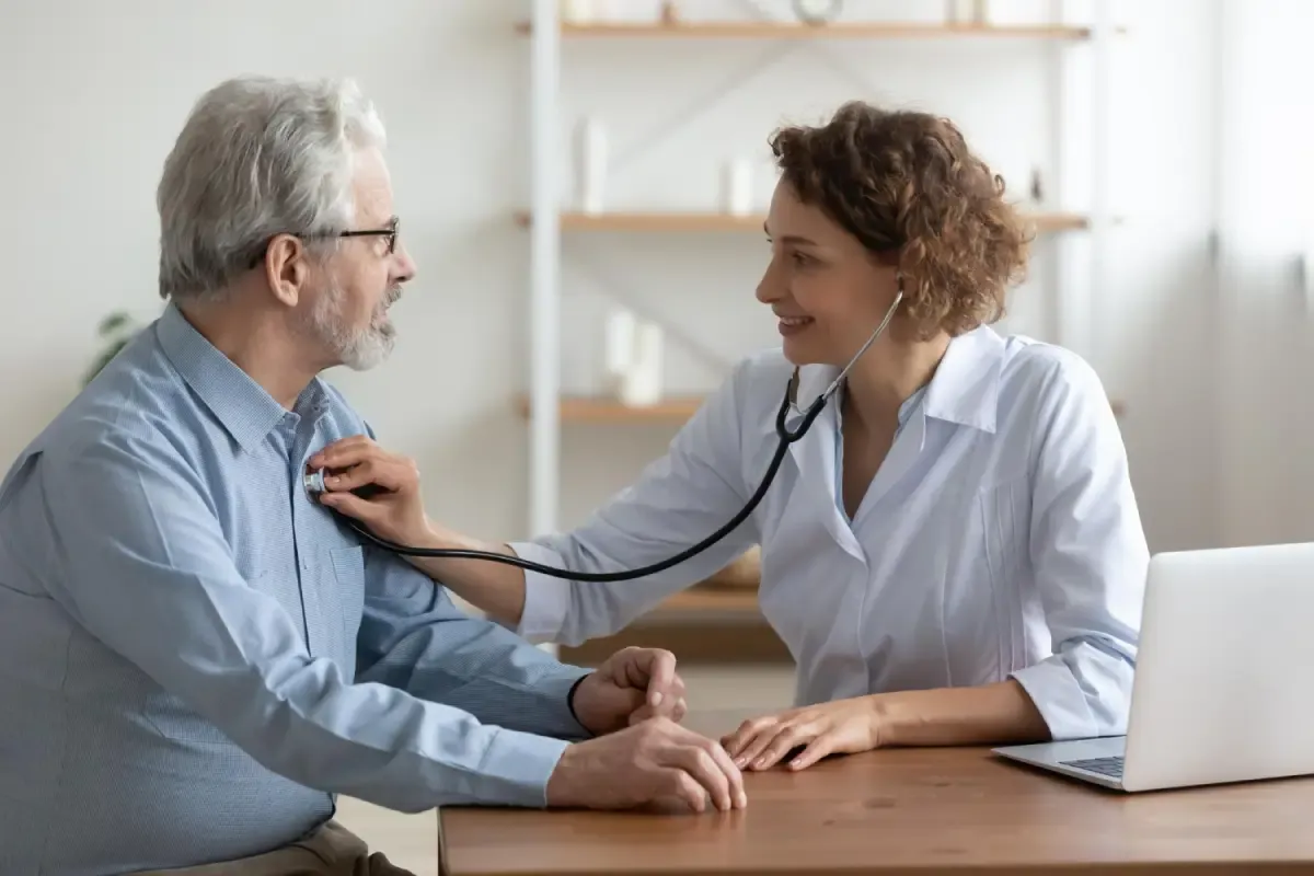 Médica que sonríe mientras usa un estetoscopio para escuchar el corazón de un hombre mayor.