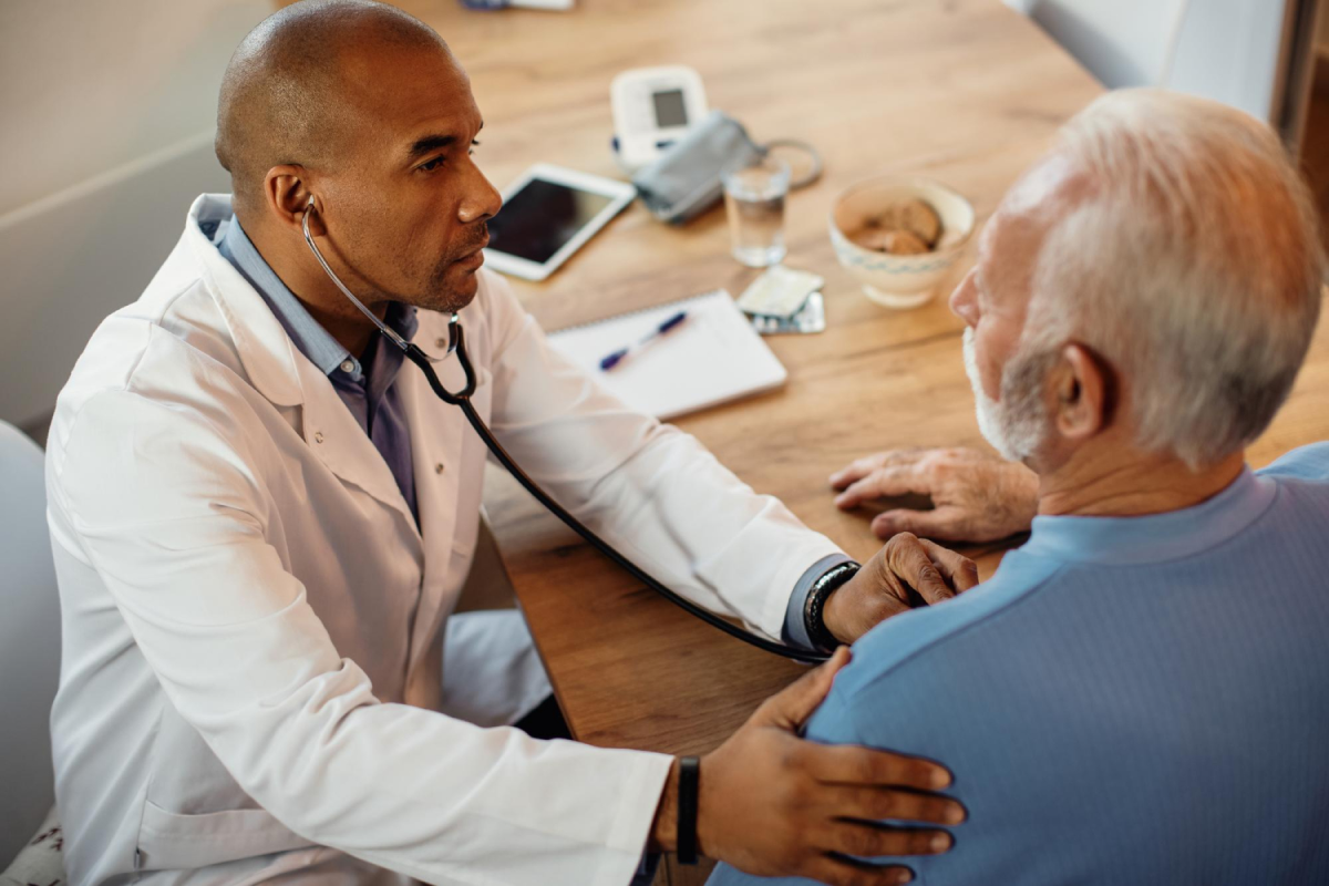 Male doctor using a stethoscope to examine an older male patient in a cozy, informal medical setting.