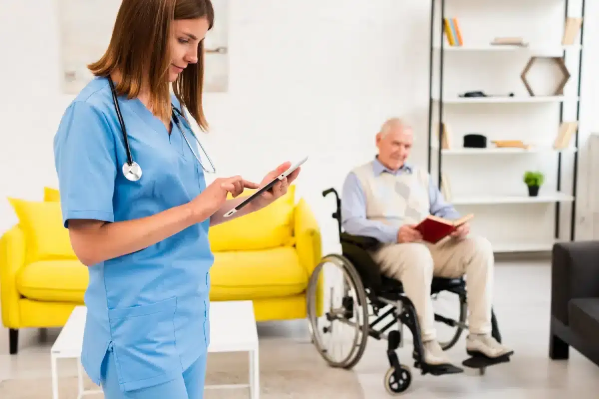 A nurse in blue scrubs using a tablet, with an elderly man in a wheelchair behind.